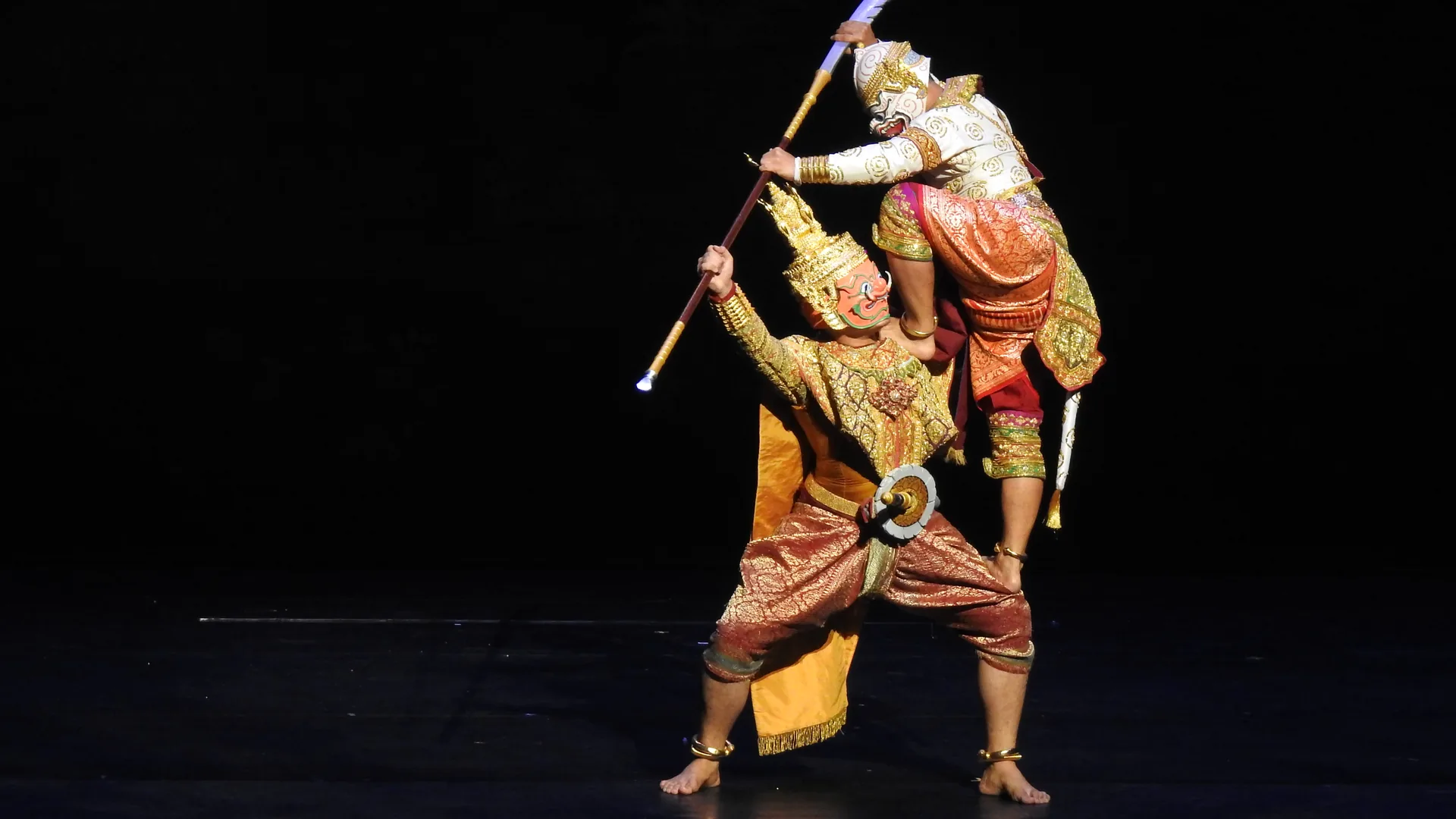 A Khon performer wearing an ornate gold and red mask and traditional costume mid-performance on a Thai stage